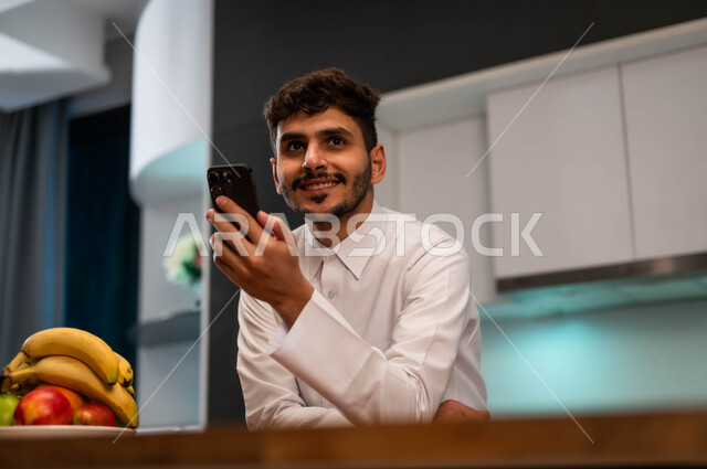 محادثات ودردشات مع الأهل و الأصدقاء باستخدام الهاتف الجوال, تصفح الانترنت ومواقع التواصل الاجماعي، رجل عربي سعودي خليجي يرتدي الزي التقليدي يقف في المطبخ أمام الطاولة ويقوم بإجراء مكالمة فيديو عبر الهاتف المحمول، التسوق الإلكتروني عن بعد، قضاء أوقات مسلية