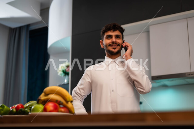 الرد على الاتصالات والمكالمات، سماع أخبار سعيدة عبر الهاتف الجوال، الدردشة مع الأهل والأصحاب، شاب عربي سعودي خليجي يرتدي الزي الرسمي ويقف أمام الطاولة في المطبخ يقوم بإجراء مكالمة هاتفية، التواصل مع الأصدقاء والاقارب باستخدام الهاتف المحمول