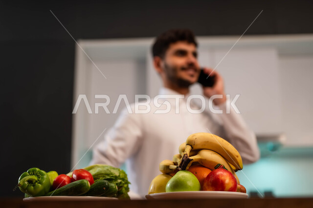 التواصل مع الأصدقاء والاقارب باستخدام الهاتف المحمول، سماع أخبار سعيدة عبر الهاتف الجوال، الدردشة مع الأهل والأصحاب، شاب عربي سعودي خليجي يرتدي الزي الرسمي ويقف أمام الطاولة في المطبخ يقوم بإجراء مكالمة هاتفية، الرد على الاتصالات والمكالمات