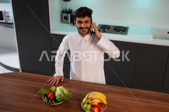 سماع أخبار سعيدة عبر الهاتف المحمول، شاب عربي سعودي خليجي يرتدي الزي الرسمي ويقف أمام الطاولة في المطبخ يقوم بإجراء مكالمة هاتفية، التواصل مع الأصدقاء والاقارب باستخدام الهاتف المحمول، الرد على الاتصالات والمكالمات، الدردشة مع الأهل والأصحاب