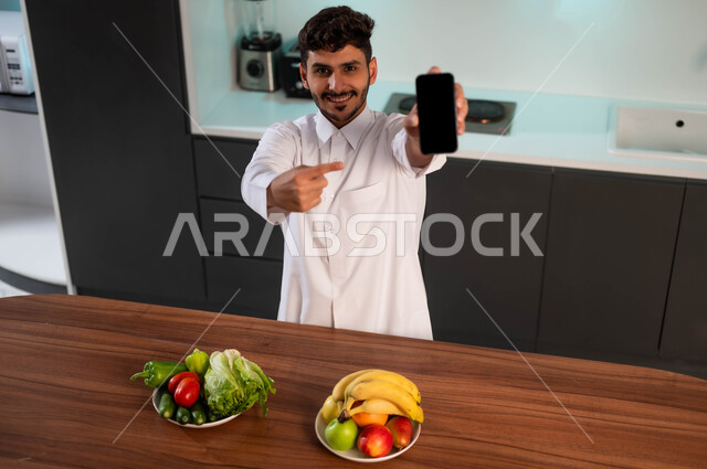 استخدام الأجهزة التقنية الحديثة، رجل عربي خليجي سعودي يرتدي الزي التقليدي يقف أمام طاولة المطبخ ويحمل بيده الهاتف المحمول بشاشة سوداء فارغة، تطور التكنولوجيا، إعداد وصفات صحية بمساعدة تطبيقات الهاتف الذكي