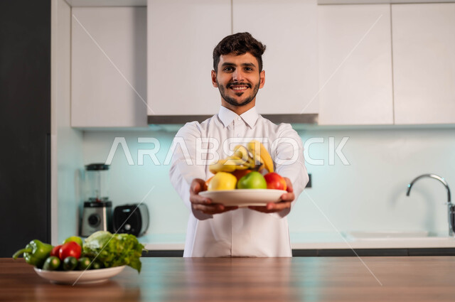 مفهوم إتباع النظام الغذائي الصحي، رجل عربي خليجي سعودي يرتدي الزي التقليدي يقف أمام طاولة المطبخ يحمل بيده طبق من الفاكهة، إيماءات السعادة والنظر إلى الكاميرا، وجبات صحية مفيدة، خضراوات طازجة