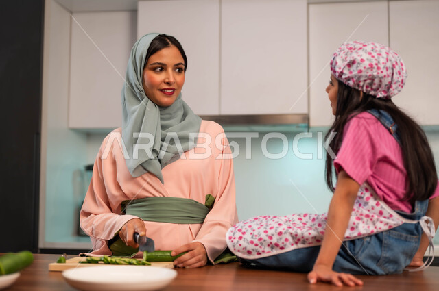 Children participating in preparing healthy food, mothers’ love and positive feelings, cutting vegetables to prepare delicious salads, the concept of healthy and beneficial eating, spending enjoyable times with the family in the kitchen, enjoying doing household chores with the daughter, a Saudi Gulf Arab mother standing with her daughter in the kitchen