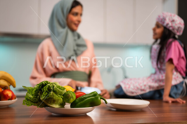 فتاه ترتدي زي الطبخ تساعد والدتها تجلس على الطاوله، مفهوم المحبة والترابط الأسري، أجواء عائلية مميزة، الاستمتاع بتحضير وصفات الطعام الصحي المنزلي، أم عربية سعودية خليجية بالزي التقليدي تعلم ابنتها أصول الطهي، مشاركة البنات مهنة الطهي