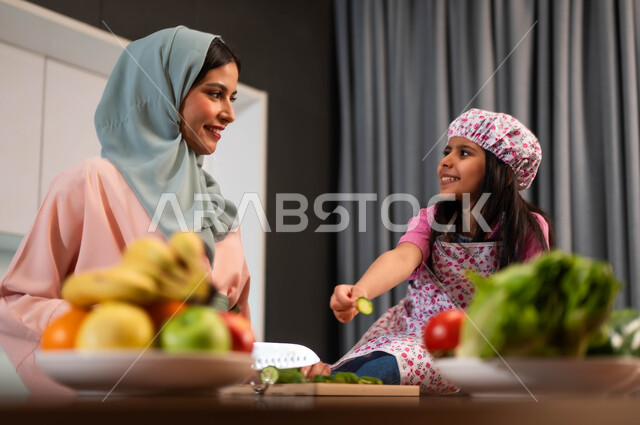 صورة مقربة أمراة عربية سعودية خليجية محجبة مبتسمة تقطع الخيار مع ابنتها في المطبخ، تجهيز الطعام وتقطيع الخضراوات، الاهتمام بالابناء ومشاركتهم بإعداد الطعام، قضاء وقت مسلي مع العائلة، تبادل الأحاديث بين الأم وابنتها في المطبخ، تحضير وجبة صحية بالخضار