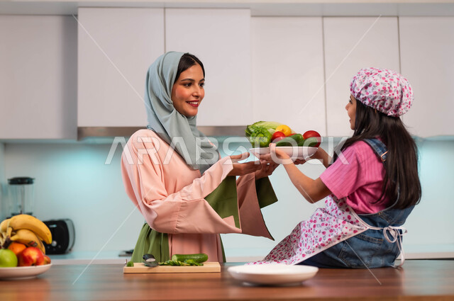 اعداد وتجهيز الطعام، تحضير وجبات صحية بالخضار الطازجة، مشاركة الاطفال في الطهي، امراة عربيه سعودية خليجية محجبه تقوم بتحضير الطعام بمساعدة ابنتها في المطبخ, ابتسامة الأم لطفلتها، ايماءات السعادة والفرح