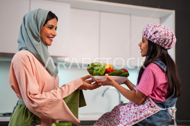 إمراة عربية سعودية خليجية محجبة ترتدي عباءة ملونة تعلم ابنتها فنون الطهي، المشاركة بالأعمال المنزلية ومهارات الطبخ، مشاركة الأطفال بالأنشطة المنزلية الترفيهية، ابتسامة الأم لطفلتها، فتاة صغيرة تقوم بمساعدة والدتها لتحضير الطعام الصحي والمفيد