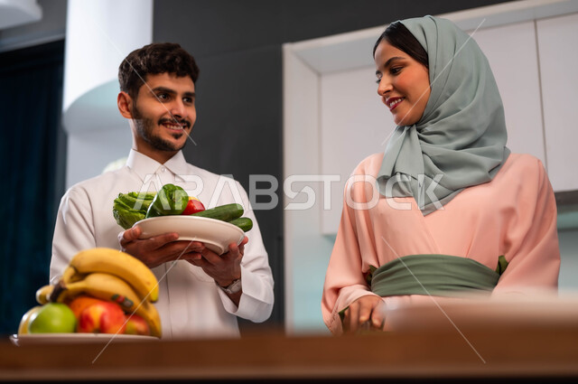 صورة مقربة لزوجان عربيان خليجيان يرتديان الزي التقليدي السعودي ويتعاونا بإعداد الطعام، تحضير وصفات الطعام الصحي والمغذي، ايماءات السعادة والامتنان، تجهيز مأكولات منزلية داخل المطبخ