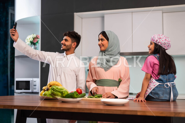 مفهوم الأسرة السعيدة، أب يلتقط صورة سيلفي عائلية جماعية تذكارية بالهاتف المحمول، قضاء أوقات عائلية ممتعة، فرحة الأبناء بقضاء وقت مسلي مع الأهل، عائلة عربية خليجية سعودية مجتمعة في المطبخ