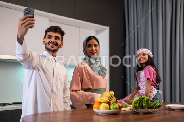 عائلة عربية خليجية سعودية مجتمعة في المطبخ، أب يقوم بالتقاط صورة سيلفي عائلية جماعية تذكارية بالهاتف المحمول، قضاء أوقات عائلية ممتعة، مفهوم الأسرة السعيدة، فرحة الأبناء بقضاء وقت مسلي مع الأهل