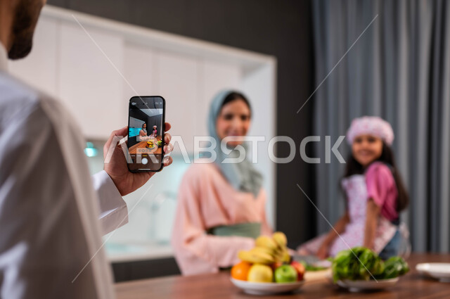 ايماءات الفرح والابتسامة والنظر إلى كاميرا الهاتف الجوال، قضاء وقت ممتع مع العائلة, أب عربي سعودي خليجي يتلتقط صورة تذكارية لعائلته من خلال الهاتف المحمول في المطبخ , اجواء اسرية مترابطة في المنزل