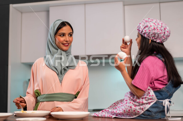 تحضير وصفات منزلية بسيطة، فتاه ترتدي زي الطبخ تساعد والدتها تجلس على الطاوله، إعداد وصفات شهية باستخدام البيض، قضاء أوقات سعيدة مع الأمهات، مشاركة البنات مهنة الطهي، امراة عربية سعودية خليجية تقوم بتحضير الطعام مع ابنتها في المطبخ