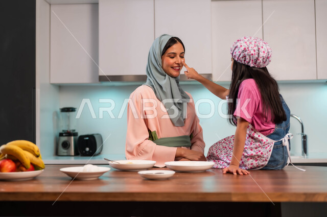 ابتسامة الأم لطفلتها، ايماءات اللعب والفرح لمشاركة البنات في الطبخ وقضاء الأوقات السعيدة في المطبخ بين الأم وابنته، إمراة عربية سعودية خليجية محجبة ترتدي عباءة ملونة تعلم ابنتها فنون الطهي، فتاه مبتسمه تقوم بمداعبة والدتها في المطبخ, طفلة أنيقة تساعد والدتها في المنزل