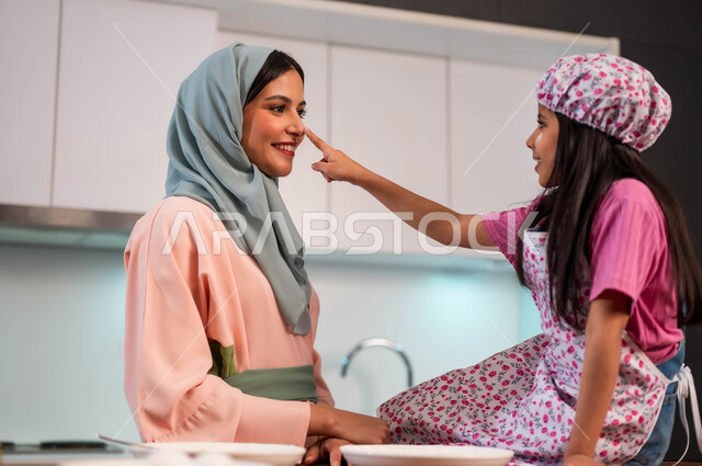 ايماءات اللعب والفرح لمشاركة البنات في الطبخ وقضاء الأوقات السعيدة في المطبخ بين الأم وابنتها ،فتاه مبتسمه تقوم بمداعبة والدتها في المطبخ, طفلة أنيقة تساعد والدتها في المنزل، إمراة عربية سعودية خليجية محجبة ترتدي عباءة ملونة تعلم ابنتها فنون الطهي ، ابتسامة الأم لطفلتها