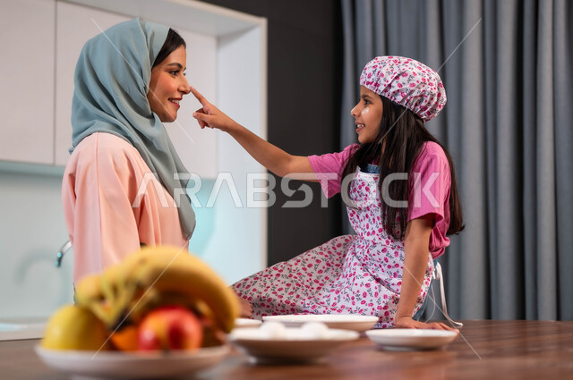 فتاه مبتسمه تقوم بمداعبة والدتها في المطبخ, ايماءات الفرح لمشاركة البنات في الطبخ، طفلة عربية سعودية خليجية ترتدي قبعة الشيف وتعلو وجهها الابتسامة مع والدتها يحضران الطعام في أجواء أسرية سعيدة وفرحة، تعليم فنون الطهي، المشاركة بالأعمال المنزلية ، تعلم وصفات جديدة