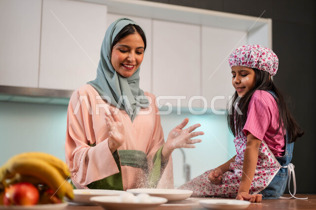 تعليم أساسيات الطبخ من أم لطفلتها ، إعداد وصفات الطبخ بأجواء عائلية مميزة ، ابتسامة إمراة عربية سعودية خليجية وهية بجانب ابنتها في المطبخ ، الشيف الصغيرة الأنيقة ، طفلة ترتدي قبعة ومئزر المطبخ ، إعداد المخبوزات وتحضير العجين