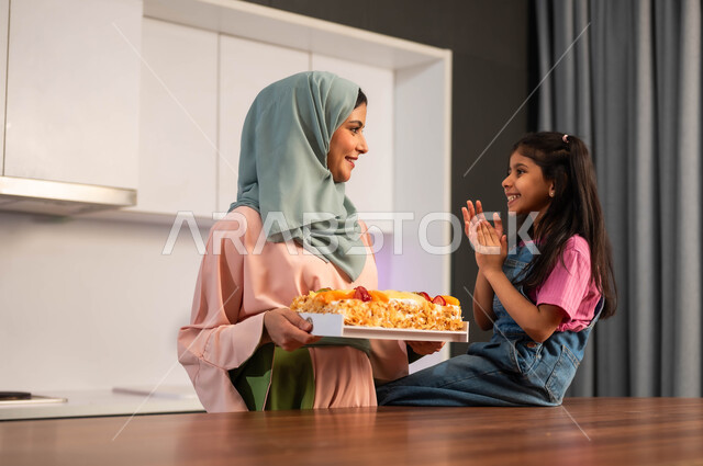 ام عربيه سعوديه خليجيه محجبه بيدها قالب من الكعك المنزلي اللذيذ , فتاه تقوم بالتصفيق لوالدتها وشكرها على اعداد الحلوى,  اعداد وتحضير الحلويات الطازجه في البيت, تقديم كيكه مزينه بالفواكه, قضاء أوقات ممتعه مع الابنه في المطبخ, ايماءات الدهشه والحماس لتذوق الكعك الشهي