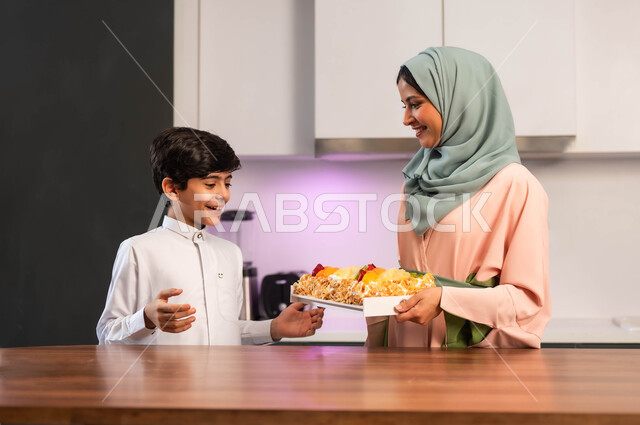 ولد متحمس لتذوق الحلوى، أم عربية خليجية سعودية تقف داخل المطبخ تحمل بيدها قالب الكيك، ايماءات الحماس والفرح, قضاء أوقات ممتعة مع الأبناء، مهارات وفنون الطبخ, إعداد وتحضير الحلويات بمكونات طبيعية طازجة