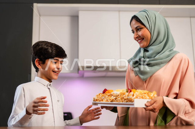 قضاء أوقات ممتعة مع الأبناء، مهارات وفنون الطبخ, ايماءات السعادة والسرور بين افراد العائلة, أم عربية خليجية سعودية تقف داخل المطبخ تحمل بيدها قالب الكيك، ولد متحمس لتذوق الحلوى، إعداد وتحضير الحلويات بمكونات طبيعية طازجة