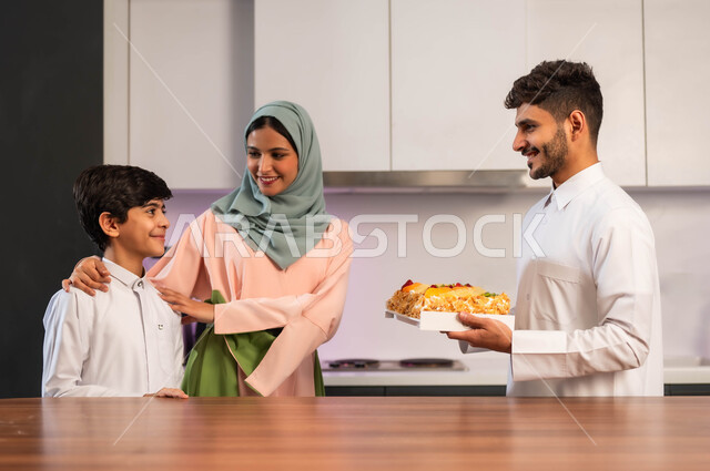 إعداد وتحضير الحلويات بمكونات طبيعية طازجة، مهارات وفنون الطبخ, قضاء أوقات ممتعة مع الأبناء، ايماءات السعادة والسرور بين افراد العائلة, أب عربي خليجي سعودي يقف داخل المطبخ يحمل بيده قالب الكيك، أم تقف بجانب ولدها