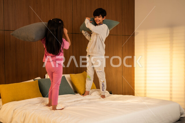 نشاطات ترفيهية مسلية، قضاء أوقات عائلية ممتعة، ولد وفتاة عربيان خليجيان سعوديان مبتسمان يلعبان على السرير يمسكان بأيديهما الوسائد، إيماءات السعادة والسرور والمرح، مفهوم الاستمتاع باللعب بين الأخوة