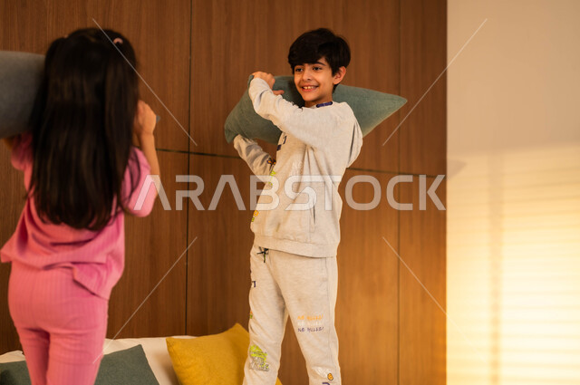أنشطة ترفيهية مسلية، قضاء أوقات عائلية ممتعة، ولد وفتاة عربيان خليجيان سعوديان مبتسمان يلعبان على السرير يمسكان بأيديهما الوسائد، إيماءات السعادة والسرور والمرح، مفهوم الاستمتاع باللعب بين الأخوة