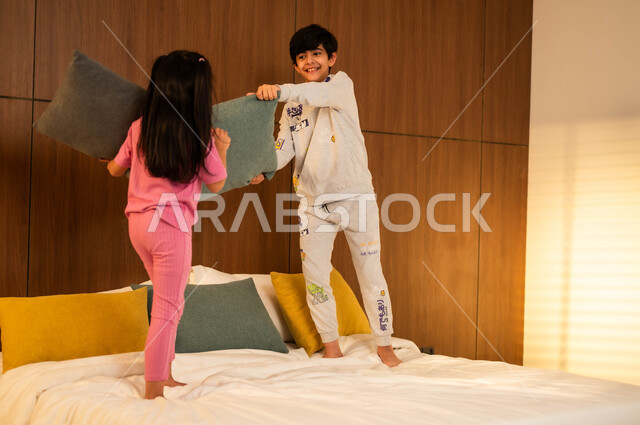 إيماءات السعادة والسرور والمرح، ولد وفتاة عربيان خليجيان سعوديان مبتسمان يلعبان على السرير يمسكان بأيديهما الوسائد، مفهوم الاستمتاع باللعب بين الأخوة، أنشطة ترفيهية مسلية, قضاء أوقات عائلية ممتعة