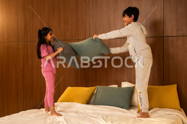 ولد وفتاة عربيان خليجيان سعوديان مبتسمان يلعبان على السرير يمسكان بأيديهما الوسائد، إيماءات السعادة والسرور والمرح، مفهوم الاستمتاع باللعب بين الأخوة، أنشطة ترفيهية مسلية, قضاء أوقات عائلية ممتعة