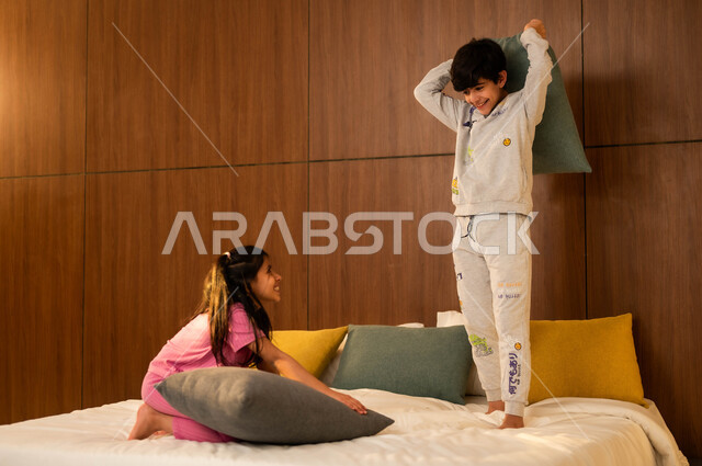 الاستمتاع في اللعب بين الأخوة، قضاء أوقات عائلية ممتعة، ولد وفتاة عربيان خليجيان سعوديان مبتسمان يلعبان على السرير يمسكان بأيديهما الوسائد، إيماءات السعادة والسرور والمرح، أنشطة ترفيهية مسلية