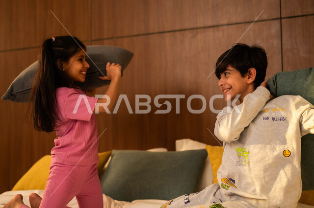 قضاء أوقات عائلية ممتعة، ولد وفتاة عربيان خليجيان سعوديان مبتسمان يلعبان على السرير يمسكان بأيديهما الوسائد، إيماءات السعادة والسرور والمرح، مفهوم الاستمتاع باللعب بين الأخوة، أنشطة ترفيهية مسلية