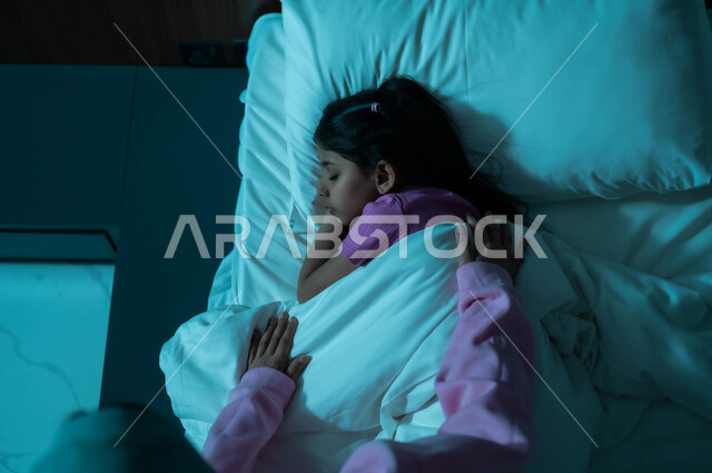 فوائد النوم المبكر، مفهوم العناية والاهتمام بالأولاد، صورة من الأعلى ليد أم عربية خليجية سعودية تطمئن على ابنتها قبل النوم، طفلة تنام في سرير منعش ومريح على وسادة بيضاء ناعمة، الاسترخاء والاستمتاع بالأجواء الهادئة، اتباع أسلوب حياة صحي