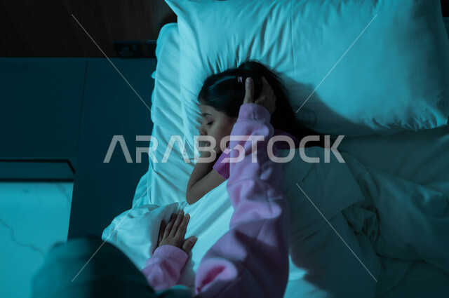مفهوم العناية والاهتمام بالأولاد، صورة من الأعلى لأم عربية خليجية سعودية تطمئن على ابنتها قبل النوم، طفلة تنام في سرير منعش ومريح على وسادة بيضاء ناعمة، فوائد النوم المبكر، الاسترخاء والاستمتاع بالأجواء الهادئة، اتباع أسلوب حياة صحي