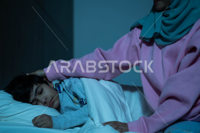 مفهوم العناية والاهتمام بالأبناء، فوائد النوم المبكر، أم عربية خليجية سعودية تطمئن على ولدها قبل النوم، طفل ينام في سرير منعش ومريح على وسادة بيضاء ناعمة، الاسترخاء والاستمتاع بالأجواء الهادئة، اتباع أسلوب حياة صحي