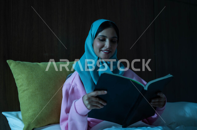 قراءة الروايات والقصص الممتعة، الشعور بالراحة والطمأنينة, سيدة عربية خليجية سعودية مبتسمة تجلس في سرير مريح تمسك بيدها الكتاب، الاسترخاء والاستمتاع بالأجواء الهادئة، اتباع أسلوب حياة صحي