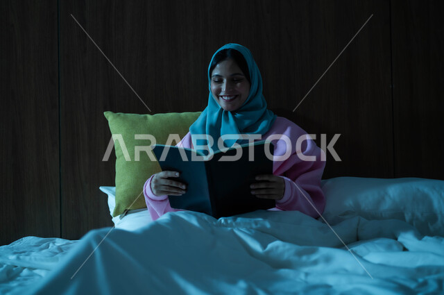 قراءة القصص والروايات الممتعة، الشعور بالراحة والطمأنينة, سيدة عربية خليجية سعودية مبتسمة تجلس في سرير مريح تمسك بيدها الكتاب، الاسترخاء والاستمتاع بالأجواء الهادئة، اتباع أسلوب حياة صحي