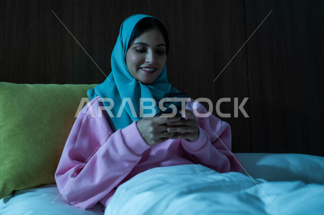 Browsing social media applications and programs, gestures of pleasure and happiness, chatting with family and friends, a smiling, veiled Saudi Arabian Gulf woman sitting in a comfortable bed, holding a mobile phone in her hand, using advanced modern technologies