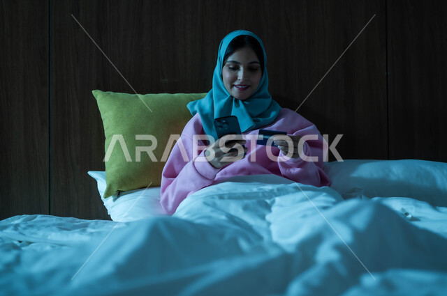 التكنولوجيا الحديثة والتقنيات المتطورة، استخدام البطاقة البنكية الالكترونية، إمرأة عربية سعودية خليجية محجبة مبتسمة تجلس على سرير مريح تعمل على الهاتف المحمول تنظر بإيماءات السعادة والسرور، مفهوم الدفع عن بعد عبر تطبيقات الجوال