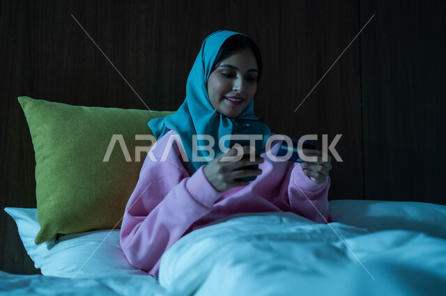 Advanced technologies and modern technology, the use of electronic bank cards, a smiling veiled Saudi Arabian Gulf woman sitting on a comfortable bed, working on a mobile phone, looking with gestures of happiness and pleasure, the concept of remote payment via mobile applications