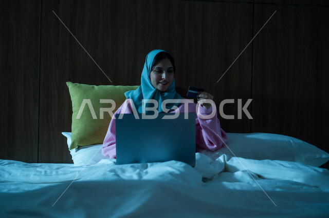 الدفع عن بعد عبر تطبيقات اللابتوب، التطور والتكنولوجيا الحديثة، استخدام البطاقة البنكية الالكترونية، إمرأة عربية سعودية خليجية محجبة مبتسمة تجلس على سرير مريح تعمل على جهاز الحاسوب المحمول تنظر بإيماءات السعادة والسرور