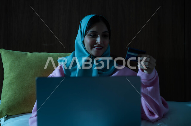 استخدام البطاقة البنكية الالكترونية، مفهوم الدفع عن بعد عبر تطبيقات اللابتوب، إمرأة عربية سعودية خليجية محجبة مبتسمة تجلس على سرير مريح تعمل على جهاز الحاسوب المحمول تنظر بإيماءات السعادة والسرور، التطور والتكنولوجيا الحديثة