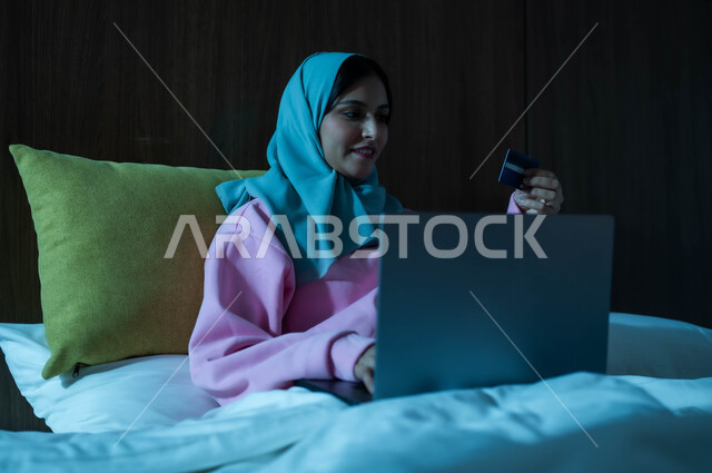 التكنولوجيا الحديثة المتطورة، إمرأة عربية سعودية خليجية محجبة مبتسمة تجلس على سرير مريح تعمل على جهاز الحاسوب المحمول تنظر بإيماءات السعادة والسرور، مفهوم الدفع عن بعد عبر تطبيقات اللابتوب، استخدام البطاقة البنكية الالكترونية