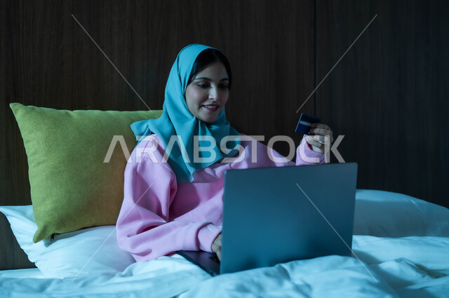 التطور والتكنولوجيا الحديثة، مفهوم الدفع عن بعد عبر تطبيقات اللابتوب، استخدام البطاقة البنكية الالكترونية، إمرأة عربية سعودية خليجية محجبة مبتسمة تجلس على سرير مريح تعمل على جهاز الحاسوب المحمول تنظر بإيماءات السعادة والسرور