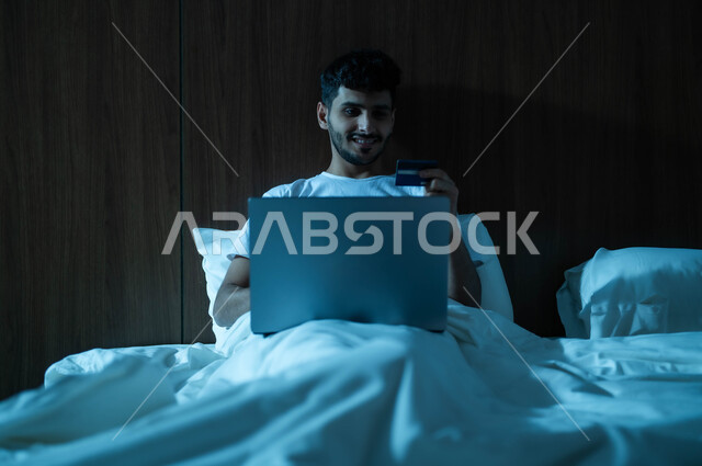 التطور والتكنولوجيا الحديثة، مفهوم الدفع عن بعد عبر تطبيقات اللابتوب، استخدام البطاقة البنكية الالكترونية، رجل عربي سعودي خليجي مبتسم يجلس على سرير مريح يعمل على جهاز الحاسوب المحمول ينظر بإيماءات السعادة والسرور