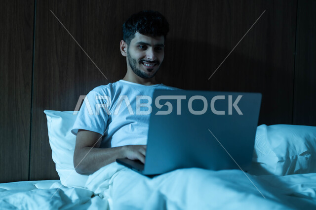 مفهوم التقنيات والتكنولوجيا الحديثة المتطورة، إنجاز مهام العمل عن بعد، رجل عربي خليجي سعودي مبتسم يجلس في سرير مريح يقوم باستخدام جهاز الحاسوب المحمول، إجراء مكالمات فيديو عبر الانترنت