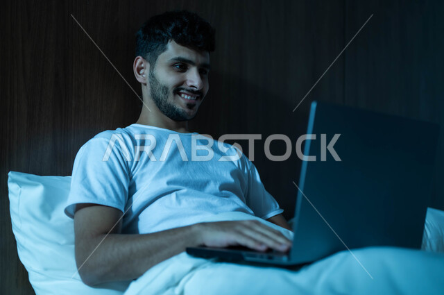 إنجاز مهام العمل عن بعد، مفهوم التقنيات والتكنولوجيا الحديثة المتطورة، رجل عربي خليجي سعودي مبتسم يجلس في سرير مريح يقوم باستخدام جهاز الحاسوب المحمول، إجراء مكالمات فيديو عبر الانترنت