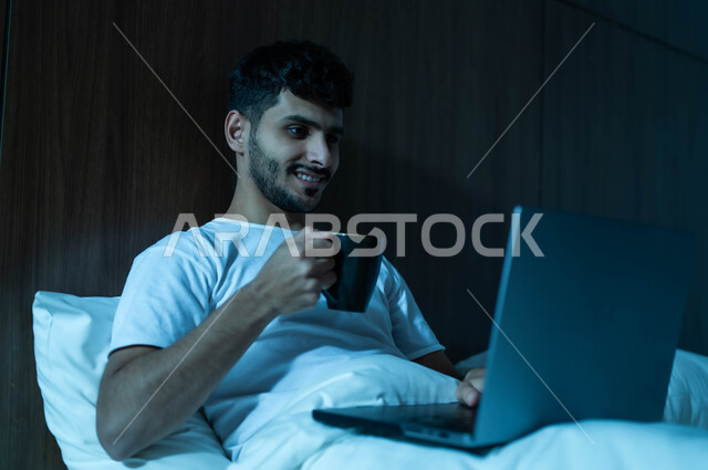 إجراء مكالمات فيديو عبر الانترنت, إنجاز مهام العمل عن بعد، مفهوم التقنيات والتكنولوجيا الحديثة المتطورة، رجل عربي خليجي سعودي مبتسم يجلس في سرير مريح يقوم باستخدام جهاز الحاسوب المحمول يحمل بيده كوب، ، تناول المشروبات الساخنة