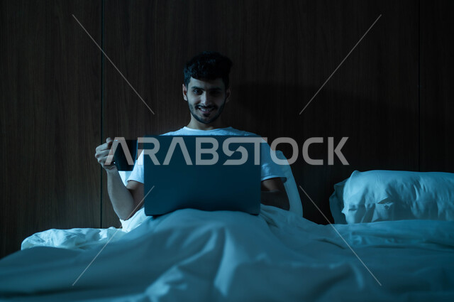 Drinking hot drinks, completing remote work tasks, the concept of advanced modern techniques and technology, a smiling Saudi Arabian Gulf man sitting in a comfortable bed using a laptop computer holding a cup in his hand, making video calls over the Internet
