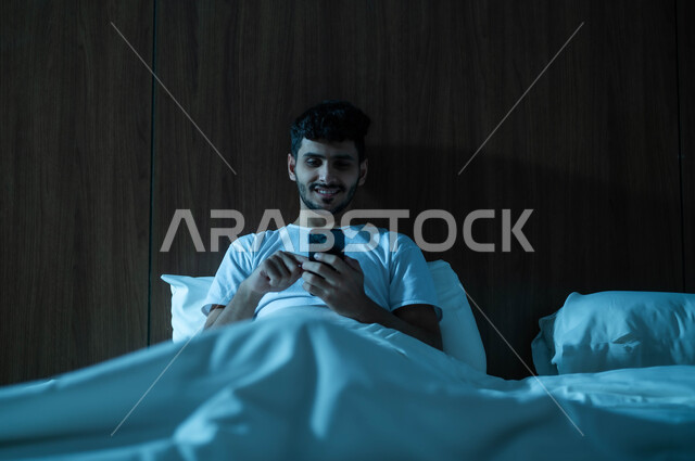 تصفح تطبيقات التواصل الاجتماعي، استخدام التقنيات والتكنولوجيا الحديثة المتطورة، رجل عربي خليجي سعودي مبتسم يجلس في سرير مريح يمسك بيده الهاتف المحمول يقوم بالتسوق الالكتروني عبر الانترنت