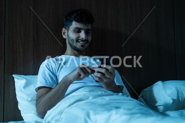 التسويق الالكتروني عبر الانترنت، استخدام التقنيات والتكنولوجيا الحديثة المتطورة، صورة مقربة لرجل عربي خليجي سعودي مبتسم يجلس في سرير مريح يمسك بيده الهاتف المحمول يقوم بتصفح تطبيقات التواصل الاجتماعي