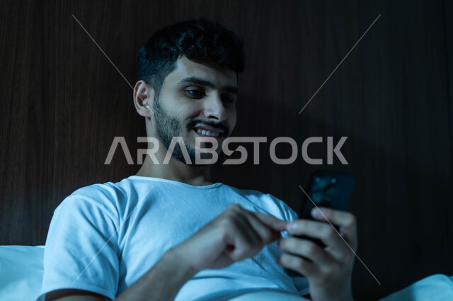 التسوق الالكتروني عبر الانترنت، استخدام التقنيات والتكنولوجيا الحديثة المتطورة، صورة مقربة لرجل عربي خليجي سعودي مبتسم يجلس في سرير مريح يمسك بيده الهاتف المحمول يقوم بتصفح تطبيقات التواصل الاجتماعي